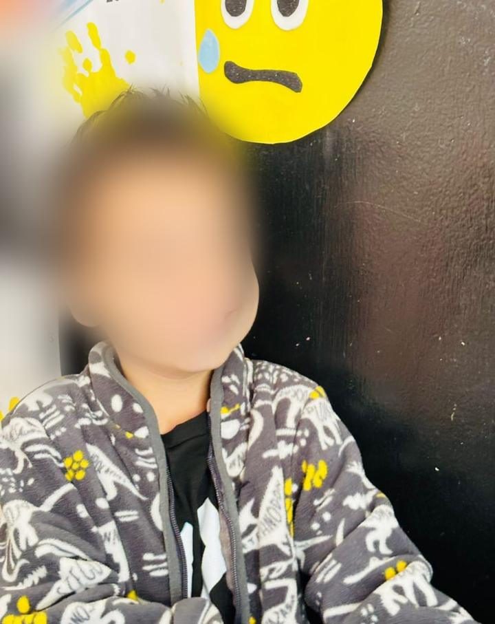 Niño con chamarra de dinosaurios frente a un pizarrón del salón; el rostro está difuminado